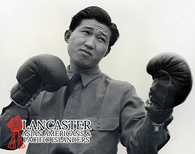 A young Asian-American man in a military uniform stands in a fighting ready position with boxing gloves on his hands.